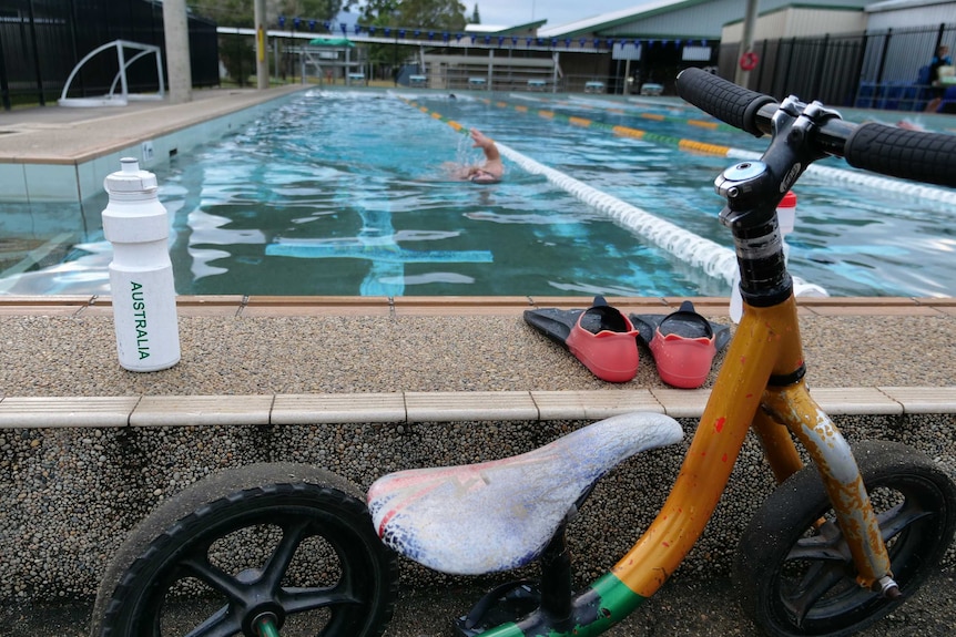 Small tricycle, flippers and 'Australia' drink bottle on the edge of the pool with backstroker approaching.