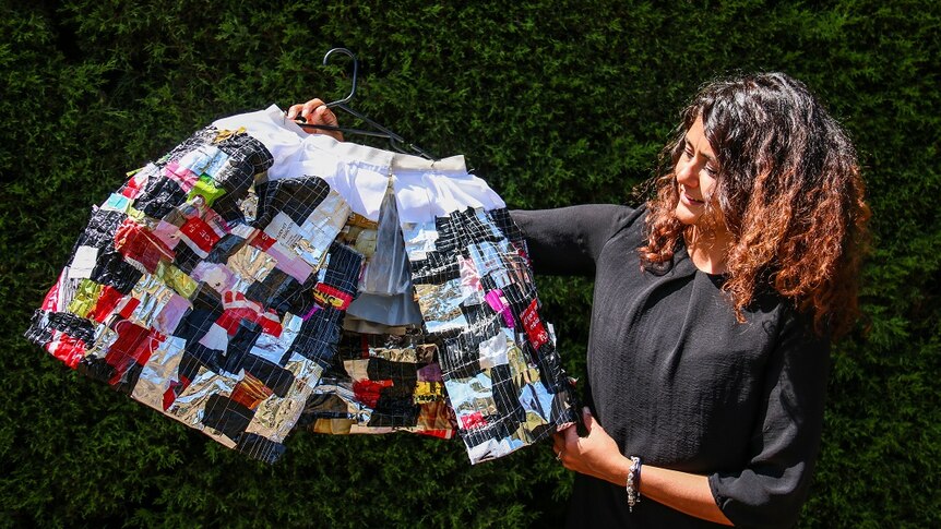 A woman holds a cape on a coat hanger made from recycled material on a sunny day in a park.