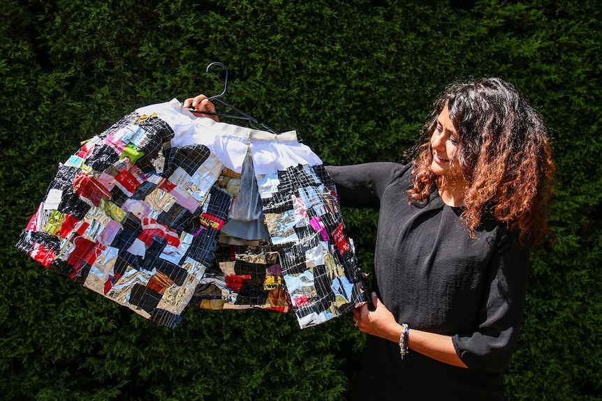 A woman holds a cape on a coat hanger made from recycled material on a sunny day in a park.