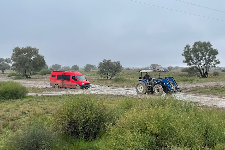 A red van being towed by blue tractor through an outback road, surrounded by grass, trees , overcast sky.
