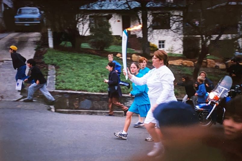 A woman carries the Sydney Olympic Torch a number of children run alongside in a suburban street.