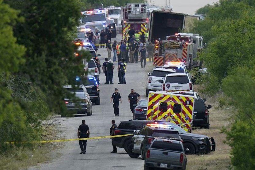 Ambulance, police and emergency vehicles and officials line a thin road with green shrub and trees alongside it 