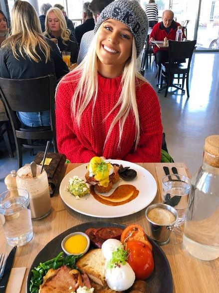 Chloe Roberts smiles in a beanie and jumper as she sits at a cafe table in front of a large serving of fritters.