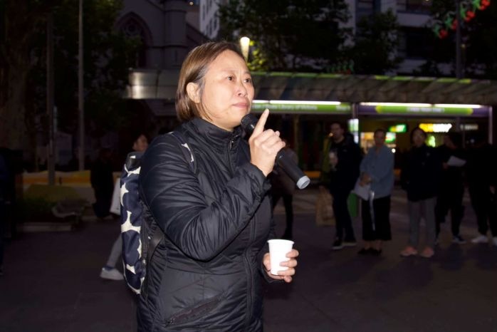 Jane Poon speaking in Melbourne at night. She is holding a microphone and standing near a tram stop.