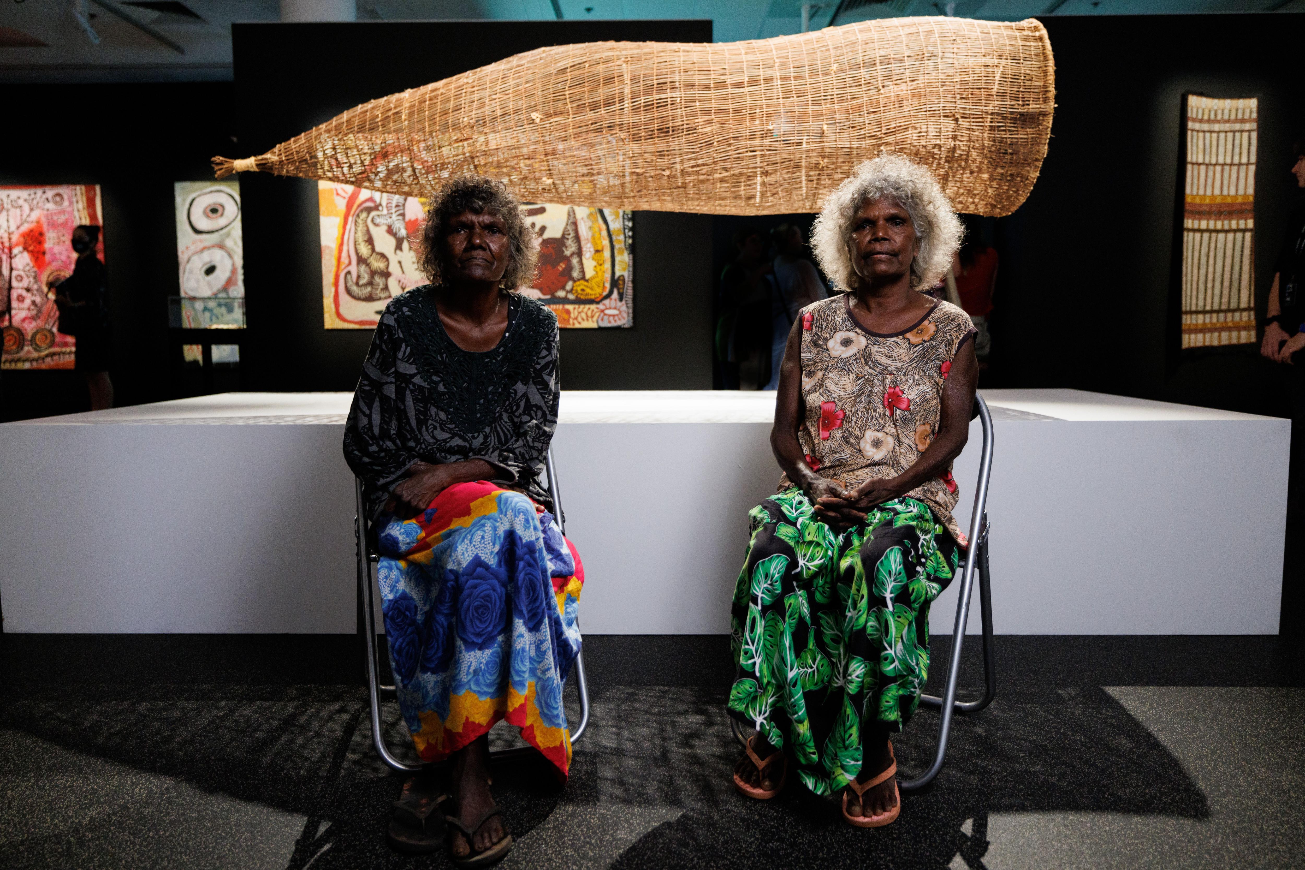 Two elderly women in an art gallery, sitting in front of a large woven fish trap suspended behind them.