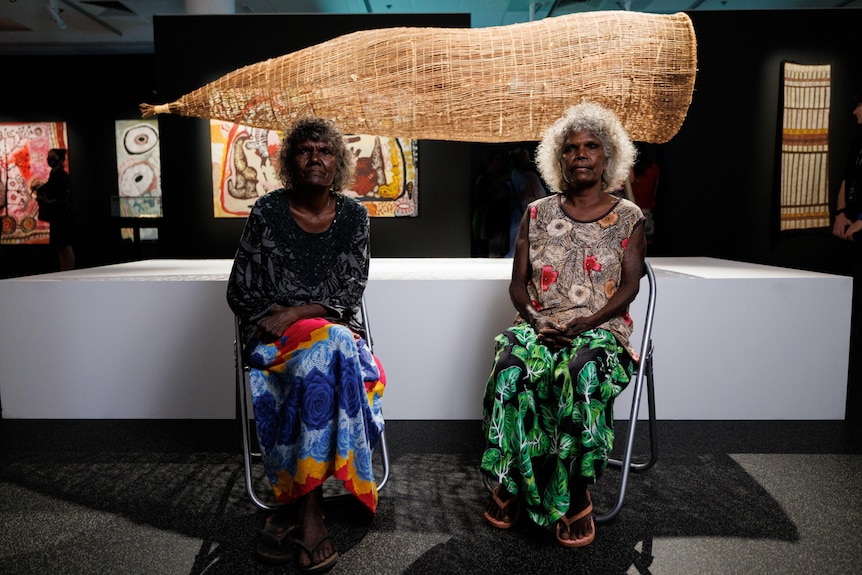 Two elderly women in an art gallery, sitting in front of a large woven fish trap suspended behind them.