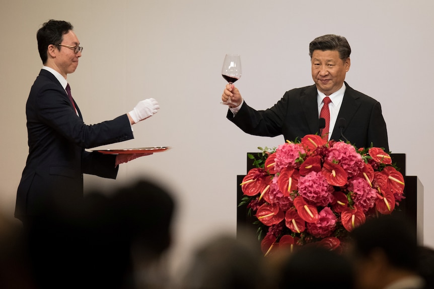 A man raising a wine glass while another man walks towards him.