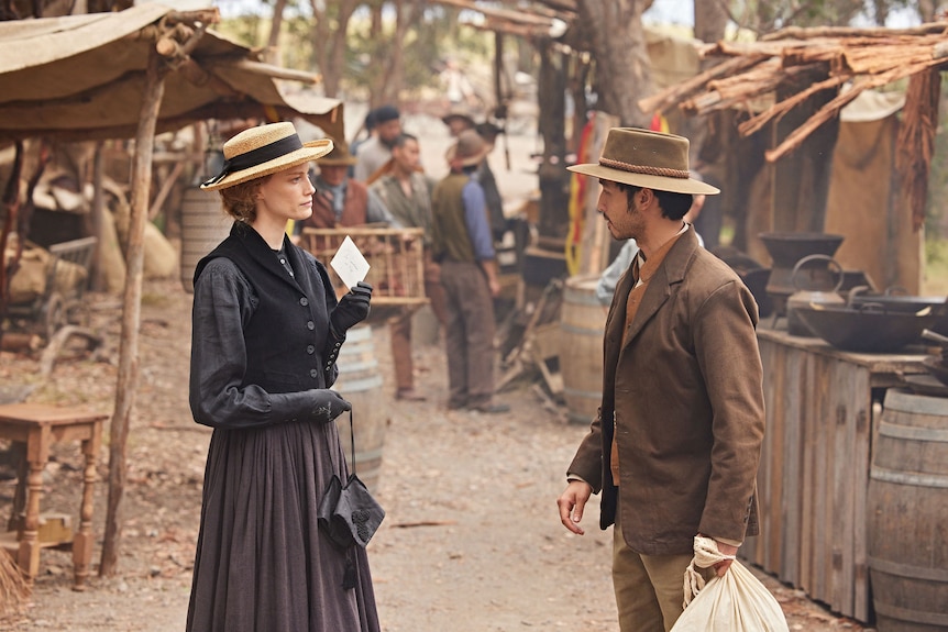 A woman and a man talk in a shanty town dressed in 1850s garb.