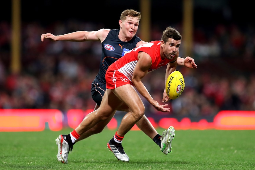 A Sydney Swans AFL player handballs in front of a GWS Giants opponent.