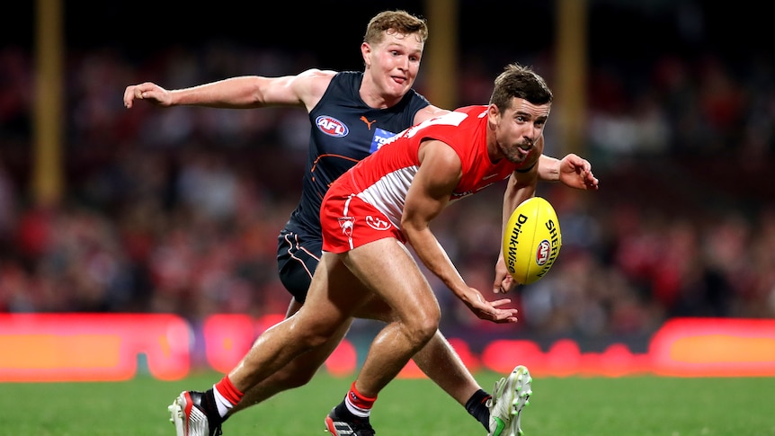 A Sydney Swans AFL player handballs in front of a GWS Giants opponent.