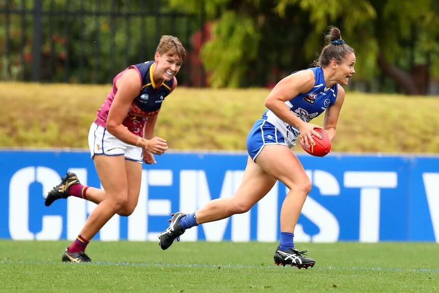 An AFLW player strains every muscle as she holds the ball and tries to outrun a defender running behind her.