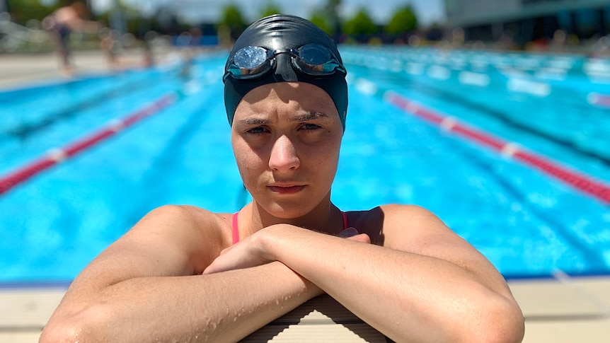 A young woman is pictured in a swimming pool, wearing a swimming cap and goggles on her head.