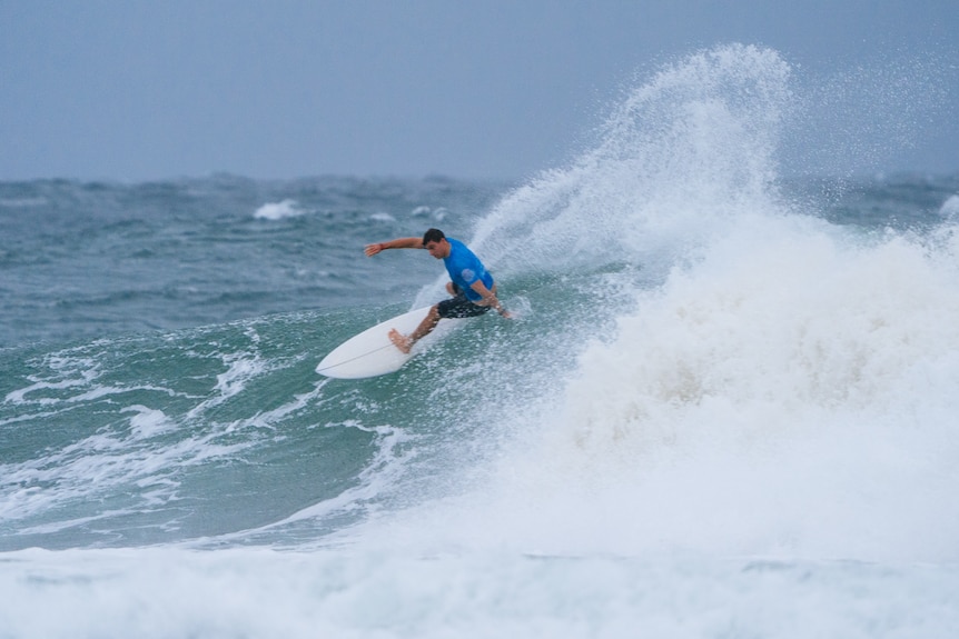 An action shot of a man wearing a blue rash shirt surfing a wave.