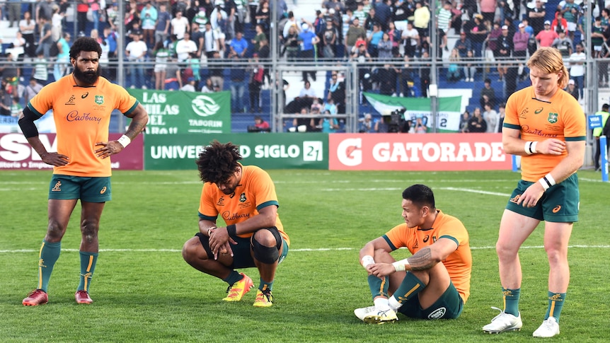 A group of Australian rugby union players look sad and serious as they stand, crouch and sit on the ground after a Test loss.
