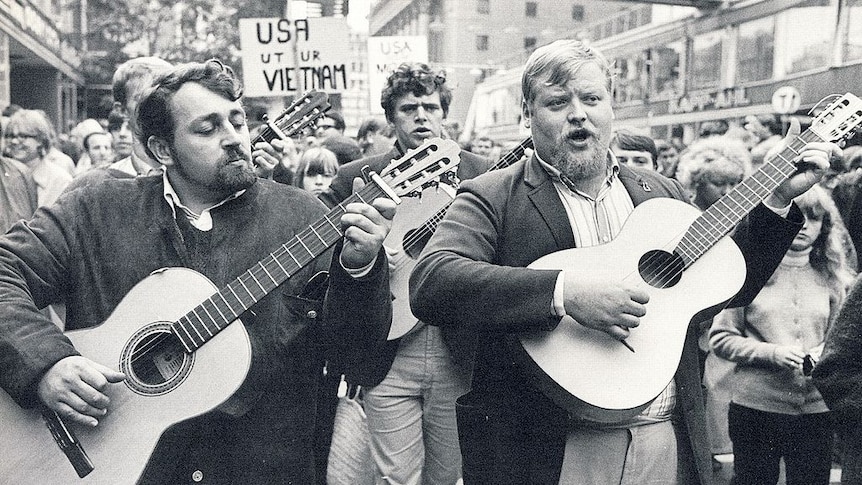 Musicians fronting a group protesters walking down street while playing guitars.