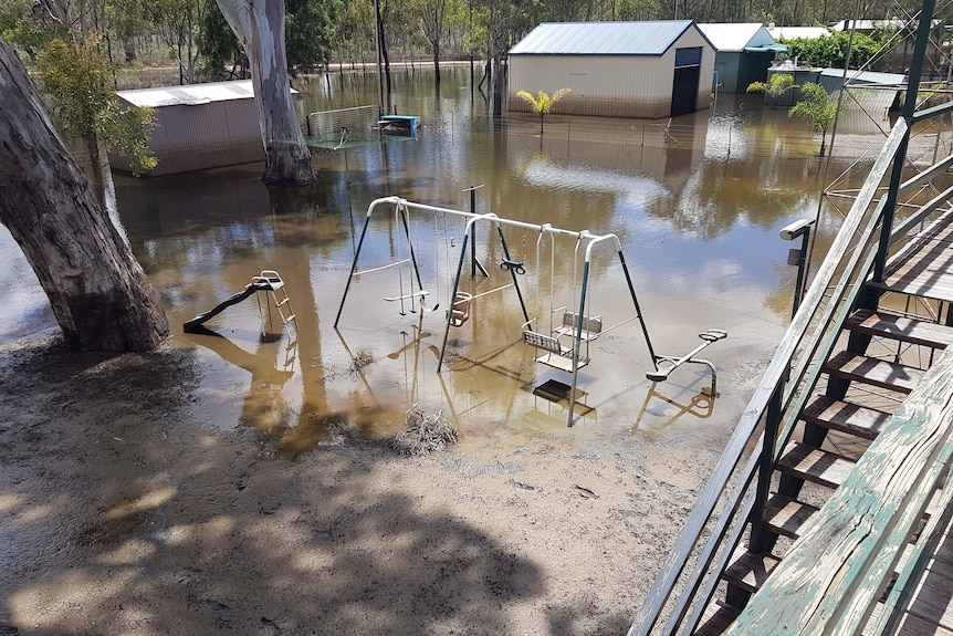 A backyard flooded, with mud and water covering part of a swing set and slide, and damaged wooden stairs.