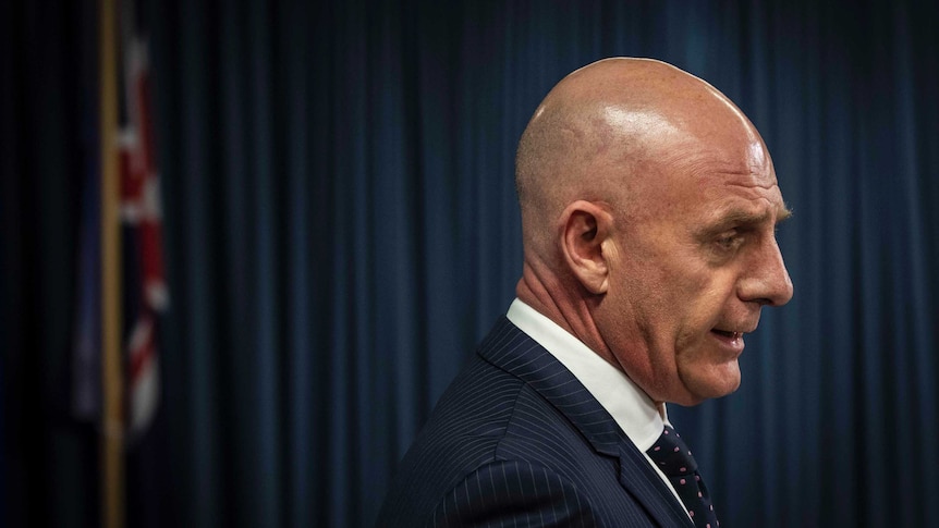 The side profile of Tasmanian Premier Peter Gutwein at a press conference with an Australian flag in the background.