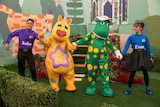 A man in a purple skivvy, a unicorn, a green dinosaur, and a woman in a blue skivvy hold hands.