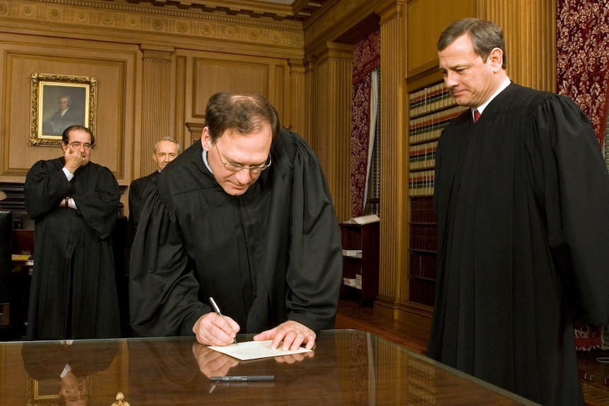 Three men in black judges' robes watch as a fourth leans down to sign a piece of paper on an ornate desk
