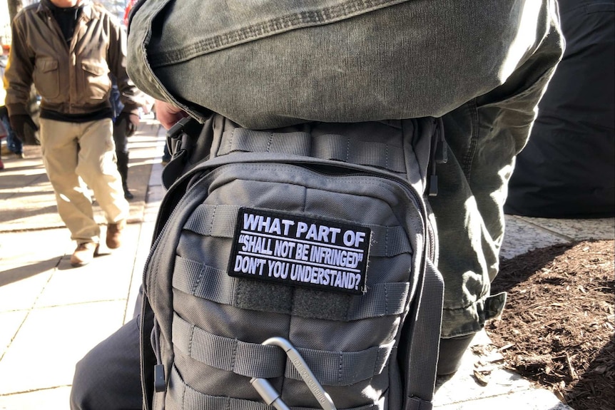 a man sits with a backpack pointed to the camera with a badge that reads what part of shall not be infringed dont you understand
