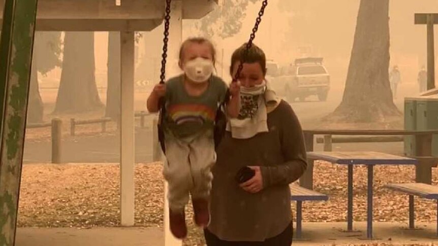A woman pushes a child on a swing, both of them wearing dust masks, as an orange haze hangs in the air in Mallacoota.