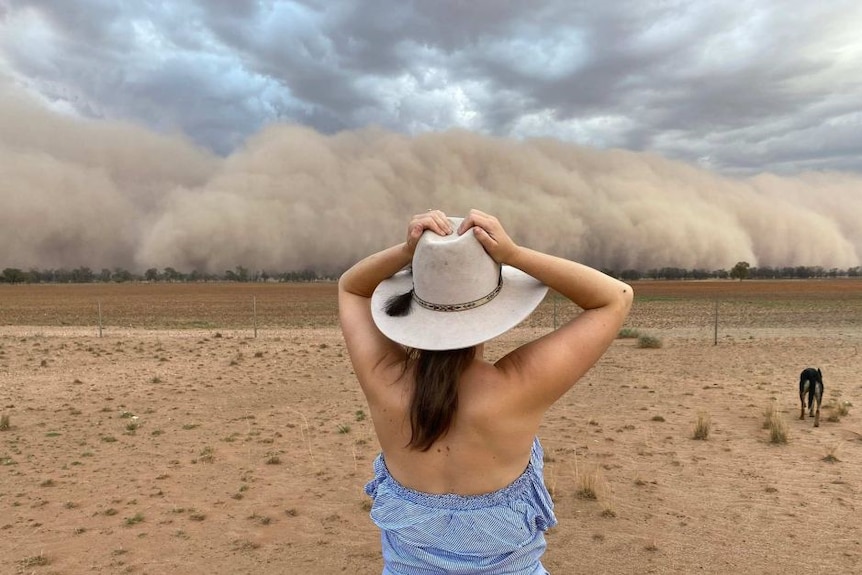A woman stands in a dry paddock with her hands on her head as a mammoth wall of dirt (a dust storm) approaches