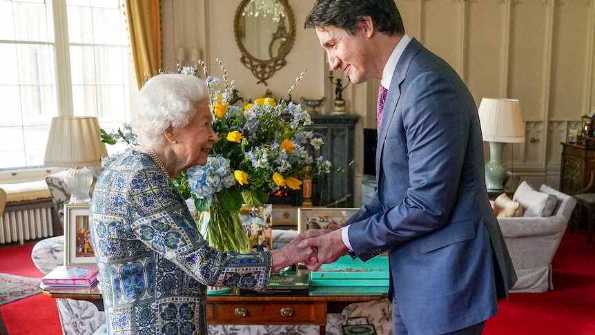 An elderly woman extends her hand to a grinning middle-aged man who holds it in both his hands in a room with red carpet.