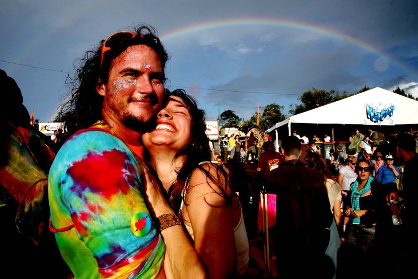 A grinning man and woman in bright clothes hug beneath a rainbow amid a crowd