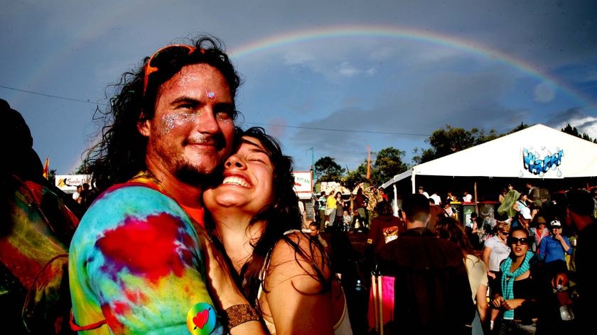A grinning man and woman in bright clothes hug beneath a rainbow amid a crowd