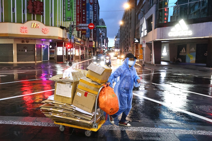A woman in a face mask and raincoat wheels a cart full of boxes through an empty street in Taipei at night