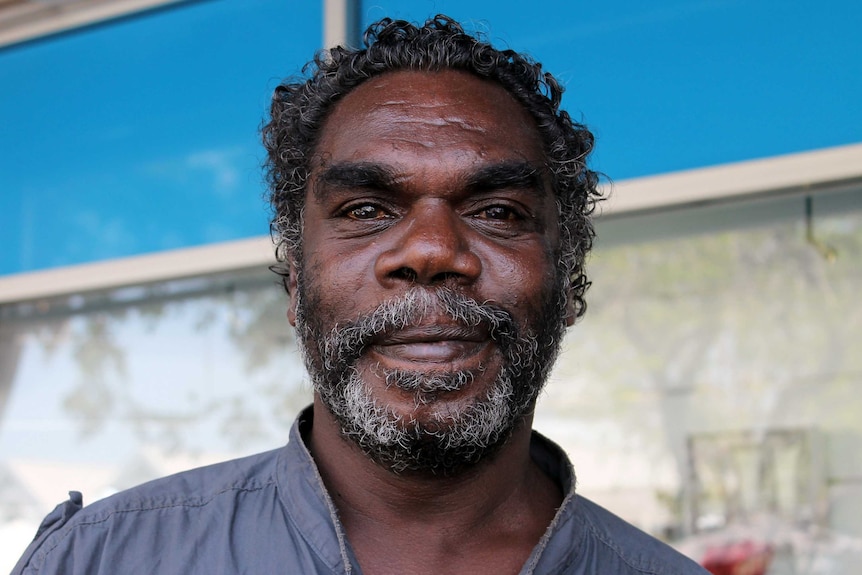 John Brown wears a grey shirt, and stands in front of a window as he looks into the camera