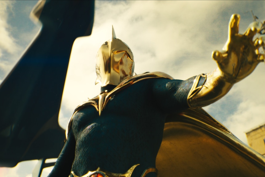 A low-angle shot of a man in a superhero costume, including a golden mask, cape and gloves