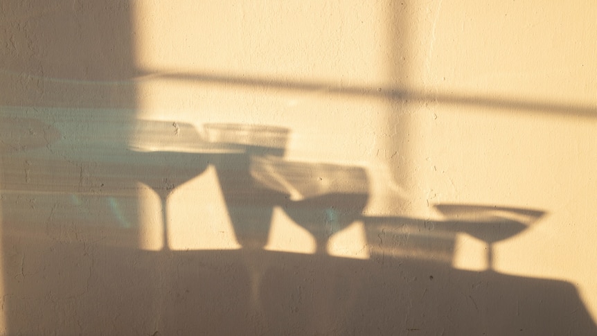 At sunset, you view a rough textured cream wall with shadows of various drink glasses spread across it.