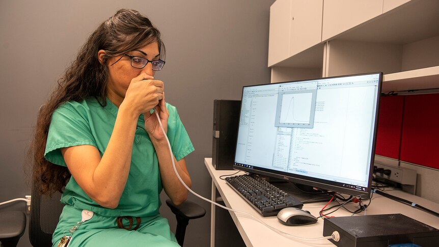 A woman in green scrubs blows into a tube that is connected to a machine plugged into a computer