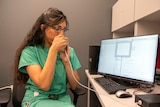 A woman in green scrubs blows into a tube that is connected to a machine plugged into a computer