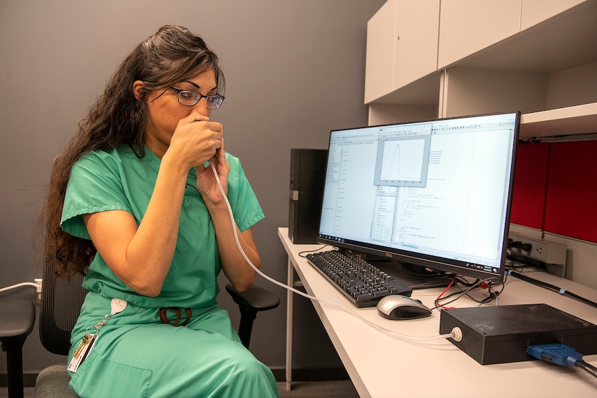 A woman in green scrubs blows into a tube that is connected to a machine plugged into a computer