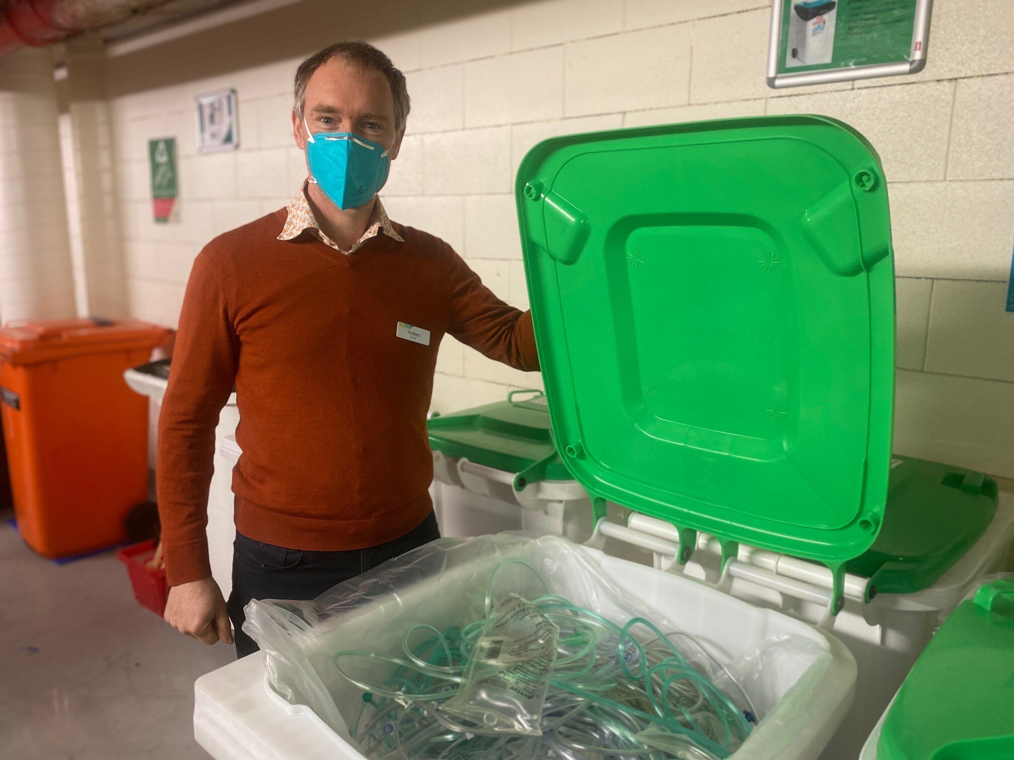 A man in a brown sweater opens a green bin with a mask on.