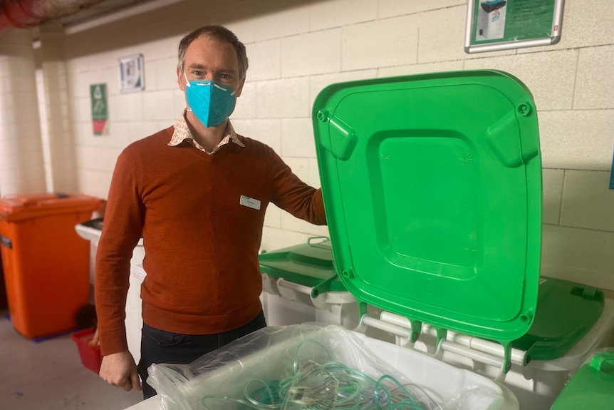 A man in a brown sweater opens a green bin with a mask on.