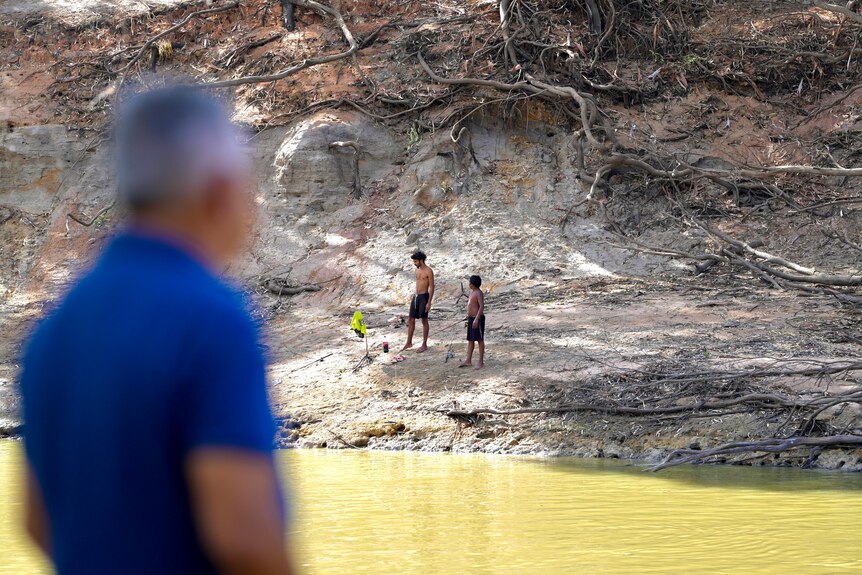 Two teenage Indigenous boys fishing in the Darling River at Wilcannia, with a man in a blue shirt watching on
