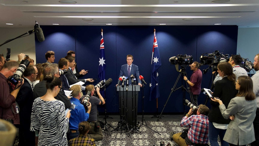 A man at a podium with microphones surrounded by journalists