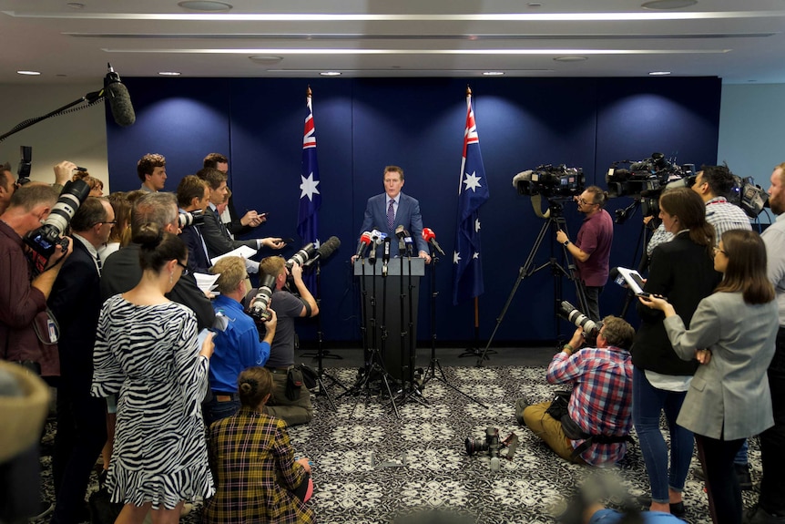 A man at a podium with microphones surrounded by journalists