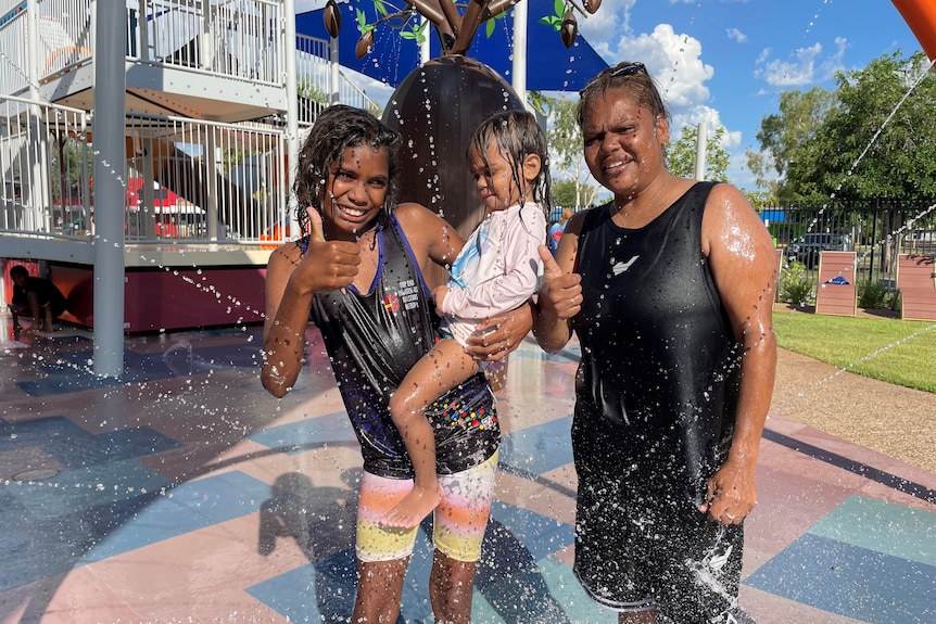 An Indigenous woman and two Indigenous children enjoying the sprinklers at a water playground