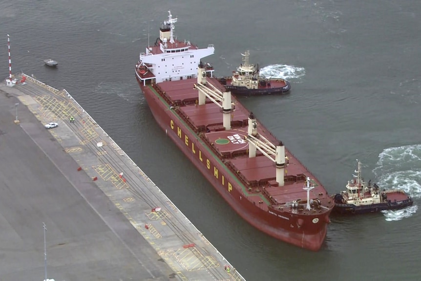A large red bulk carrier is pushed into dock by tug boats.