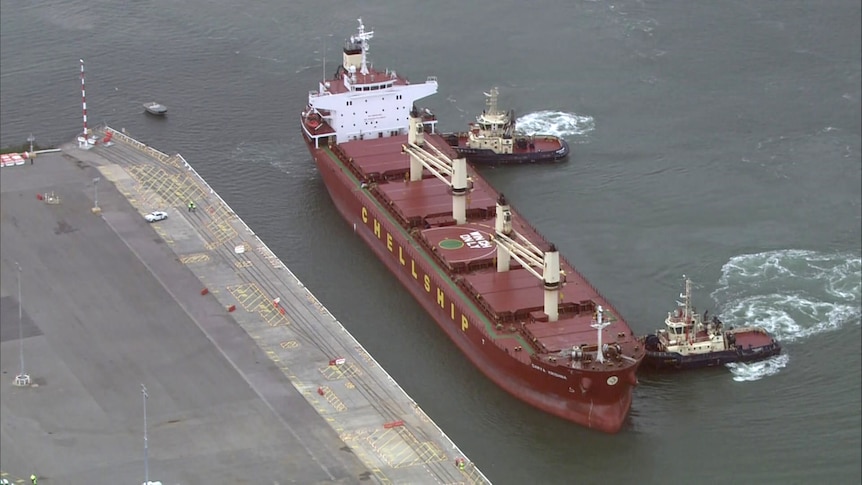 A large red bulk carrier is pushed into dock by tug boats.