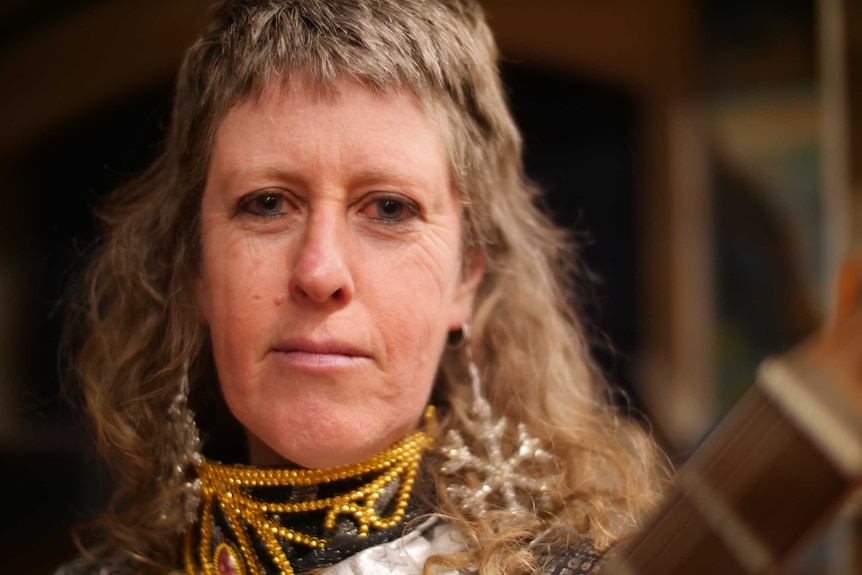 Close shot of a woman holding a guitar and wearing theatre bling around her neck looking down the lens.