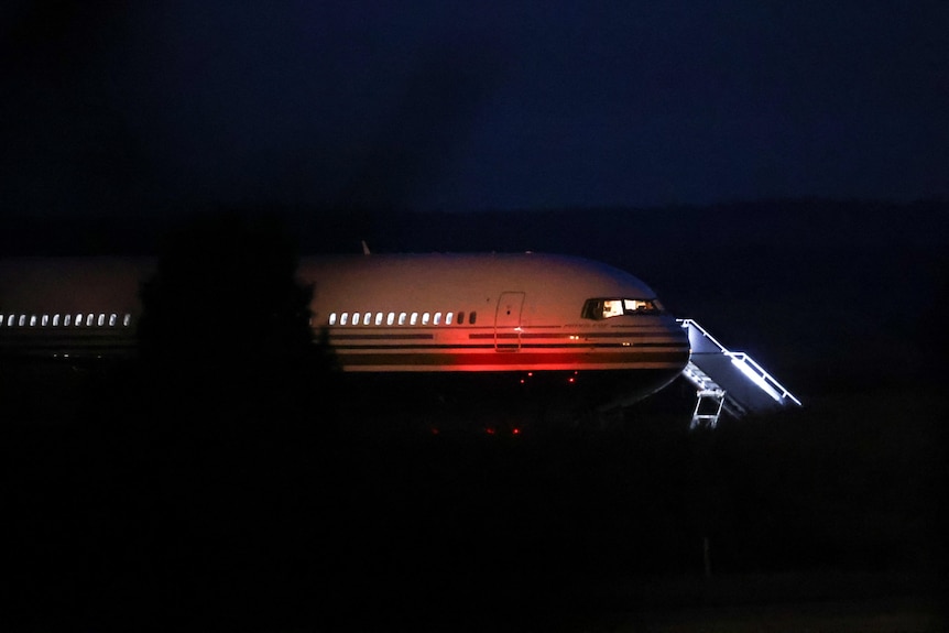 The front of a passenger plane emerges from the dark of night into lights.