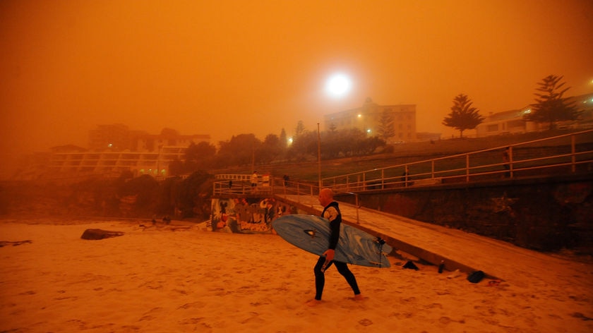 A surfer walks through a beach that is covered in dust and turning the air orange.