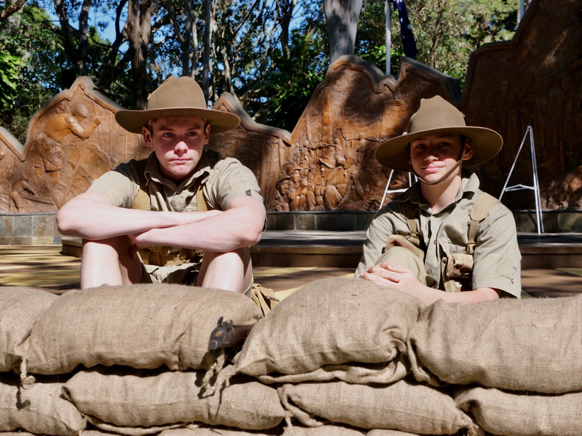 Two teenage boys sit and stare at the camera, dressed in army fatigues in front of a small wall of sandbags