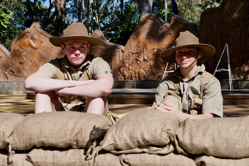 Two teenage boys sit and stare at the camera, dressed in army fatigues in front of a small wall of sandbags