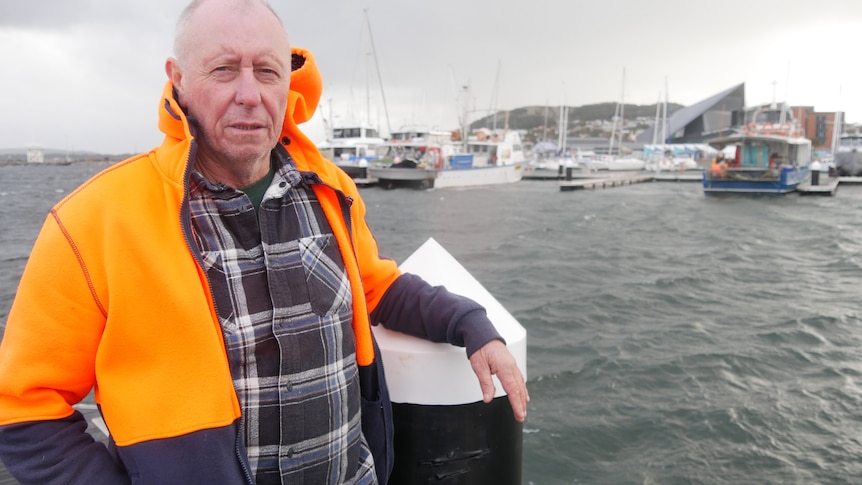 An older man in a hi-vis jacket standing on a pier in front of a hostile-looking sea.